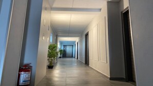 A long, narrow hallway with a fire extinguisher on the left, potted plants along the wall, and doors leading to rooms on both sides. - EM04- Studio premium - pat matrimonial (Târgu Jiu)