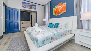 Een slaapkamer met een groot bed, een nachtkastje, een lamp, een schilderij van bloemen en een deur naar een andere kamer.