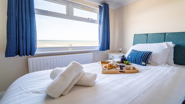 Un lit bien fait, avec un plateau contenant des croissants, une tasse de café et une petite plante. Une fenêtre offre une vue sur la plage, et la pièce présente un aménagement simple et épuré.