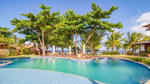Ein Schwimmbecken mit Liegestühlen und Palmen, eingebettet in eine Resortlandschaft.