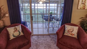 A room with a sliding glass door leading to a balcony with a table and chairs, two red armchairs with decorative pillows, and a ceiling fan.