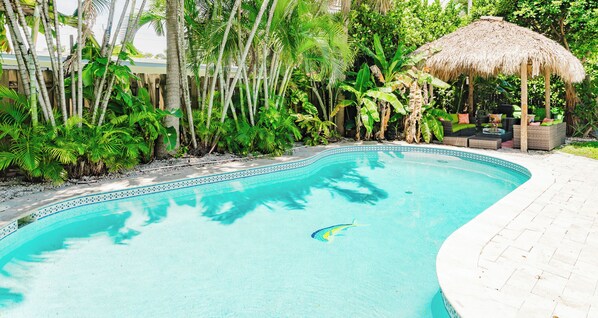 A swimming pool with a thatched roof structure and lush tropical vegetation surrounding it.