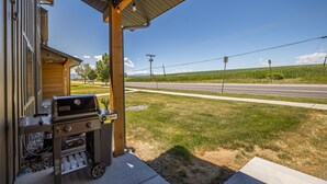 A covered outdoor area with a barbecue grill, a wooden building, and a view of a road and fields. - Brand New Townhome - 10 Minutes from Downtown (Bozeman)