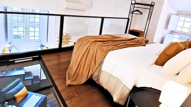 Ett modernt sovrum med ett stort fönster, en säng med ett beige täcke, ett sängbord och en hängande hylla.