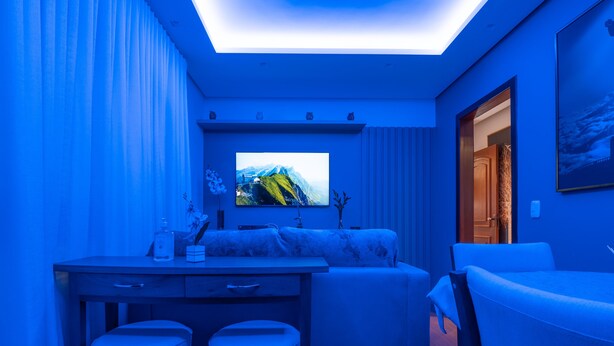 A modern room with a flat-screen TV on the wall, a sofa, a desk with two stools, and a door leading to another room. - Apartment in Gramado city center, 100m from Rua Coberta (Gramado)