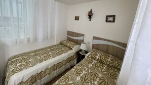 Une chambre avec deux lits simples, une fenêtre avec des rideaux légers, une lampe fixée au mur et deux tableaux encadrés.