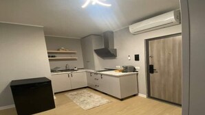 Uma cozinha compacta com fogão embutido, pia e micro-ondas. Há um cesto de lixo pequeno e uma porta à direita.