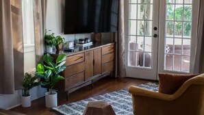 Ein Wohnzimmer mit an der Wand montiertem Fernseher, einem Holzkabinett, einem Sofa und einer zur Außenwelt führenden Tür.