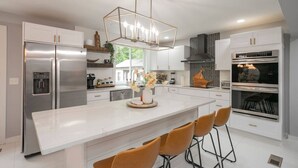 A modern kitchen with white cabinetry, stainless steel appliances, a central island with bar stools, and a decorative light fixture.