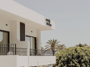 A modern building with a sign reading "Comfort Studios." It features a balcony with a metal railing, large windows, and a view of palm trees.