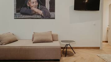 A modern living room with a beige sofa, a small round table, and a flat-screen TV mounted on the wall.