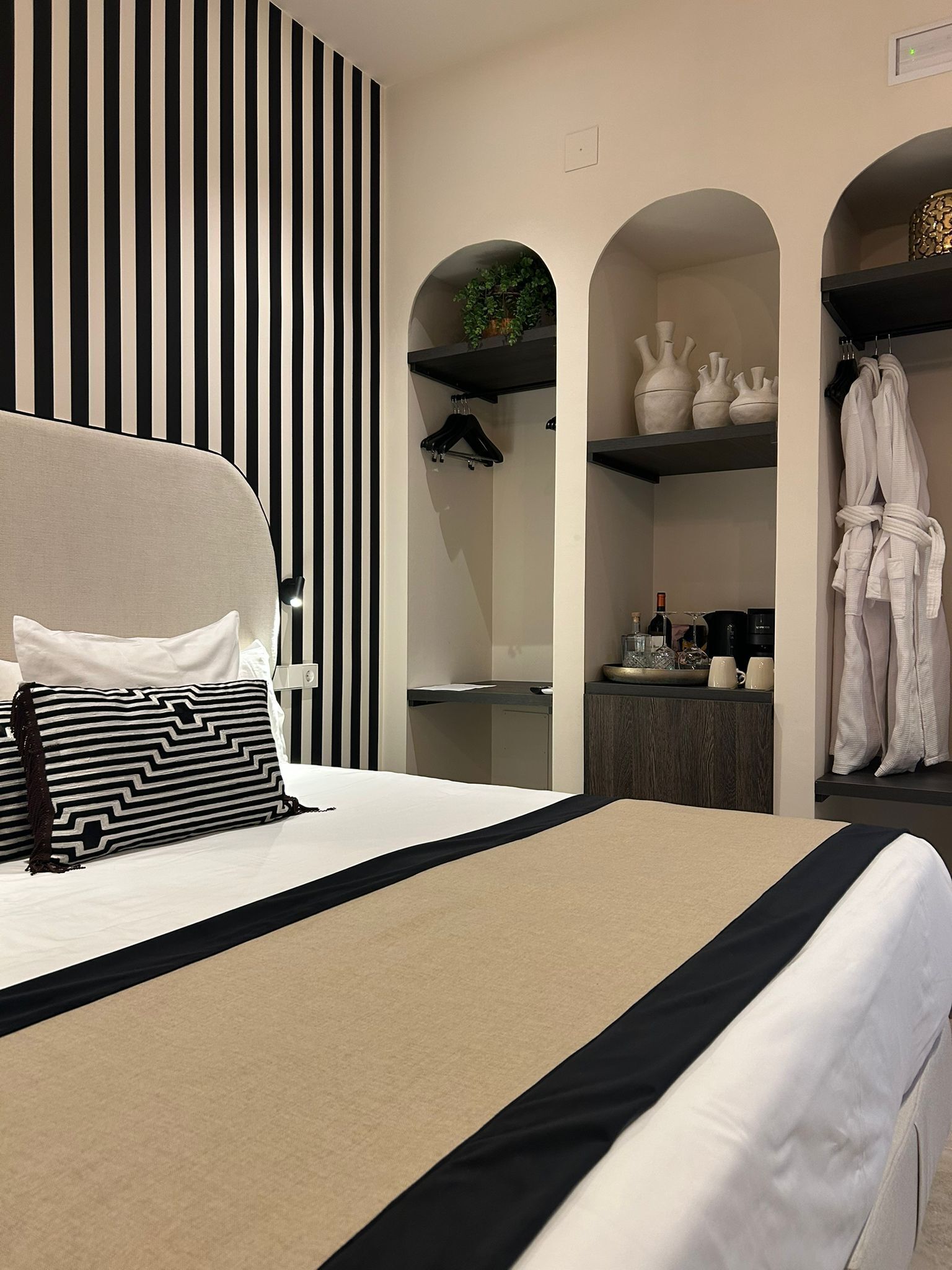 A modern hotel room with a bed, black and white striped wall, arched shelving with decorative items, and a robe rack.