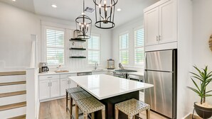 Una cocina moderna con armarios blancos, una isla central con taburetes, electrodomésticos de acero inoxidable e iluminación colgante.