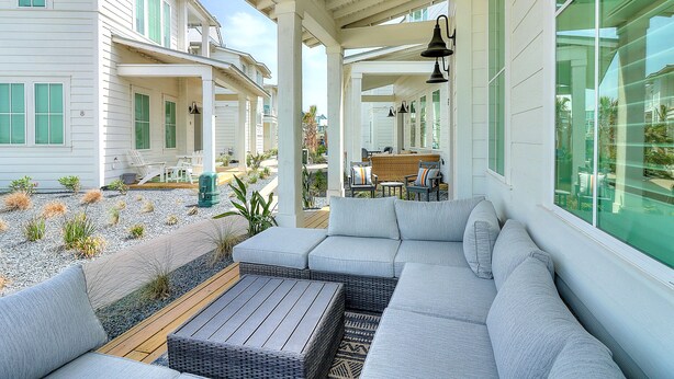 Un área de estar al aire libre con techo, que incluye un sofá y una mesa, rodeada por un patio con plantas y una terraza de madera.