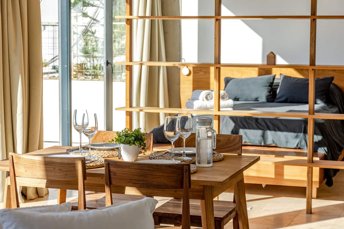 Una habitación moderna con una mesa de comedor de madera preparada para dos personas, una cama con cabecera de madera y un ventanal con cortinas.