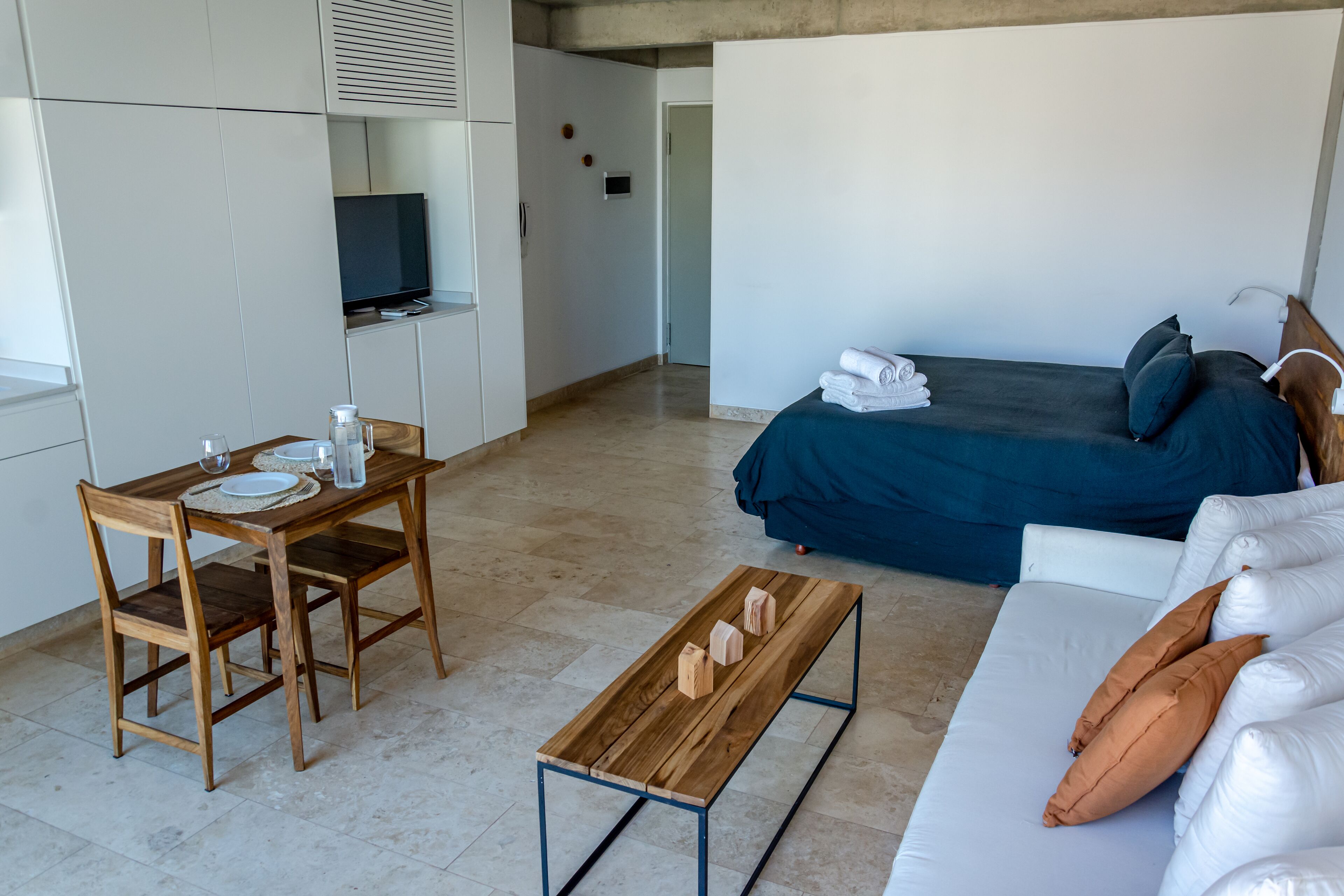 A modern hotel room with a bed, a sofa, a wooden table, and a TV.