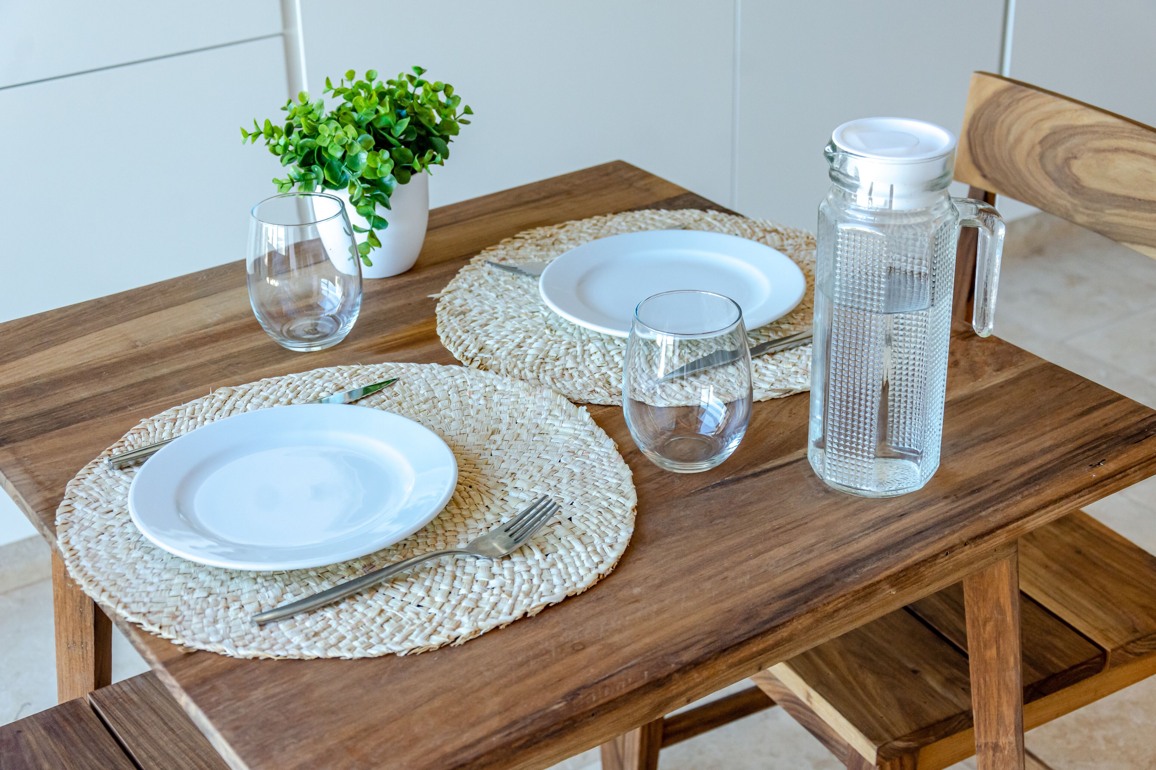 A wooden dining table set with white plates, cutlery, and a clear glass pitcher.