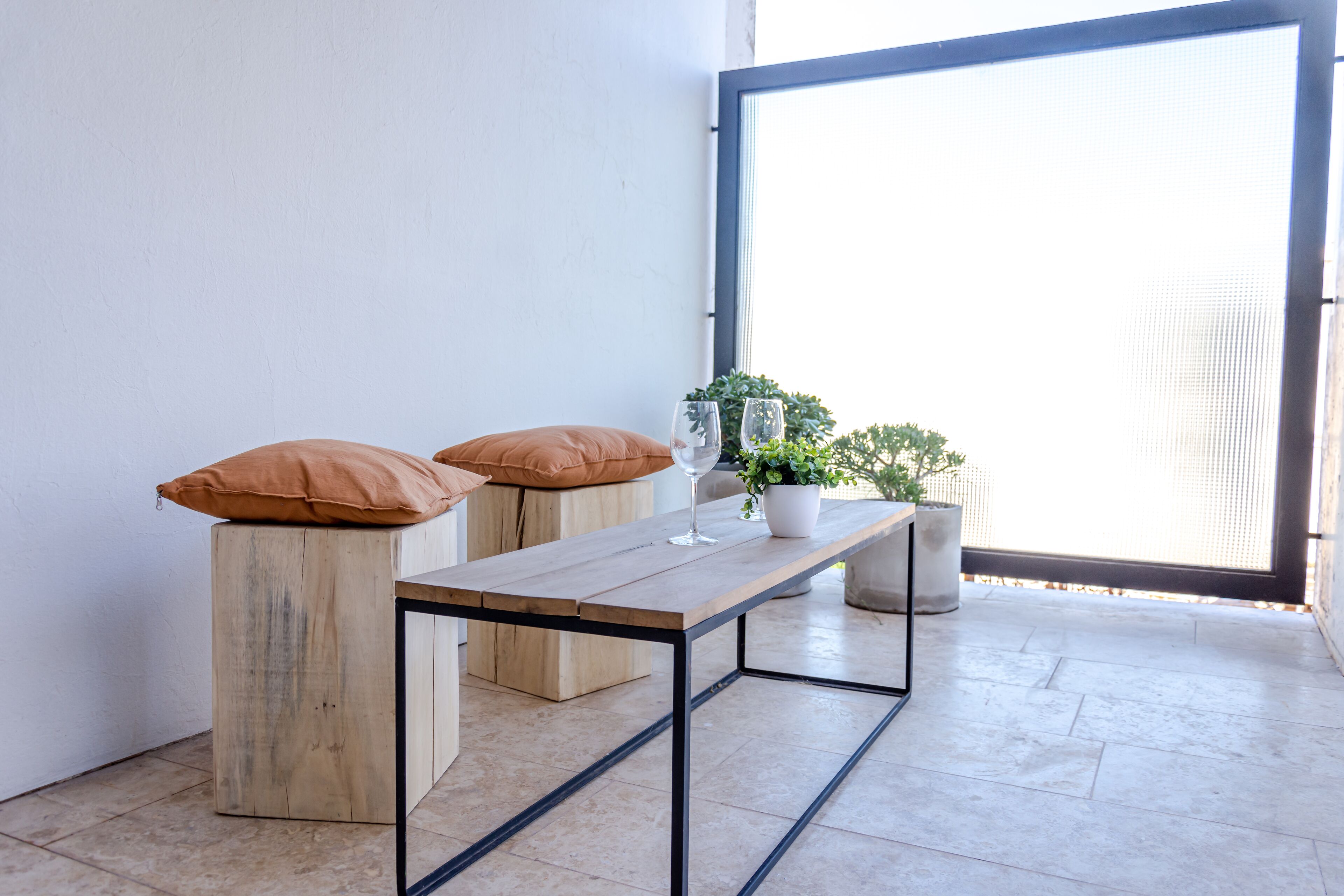 A small, modern room with a wooden table and two stools, a potted plant, and a large window.