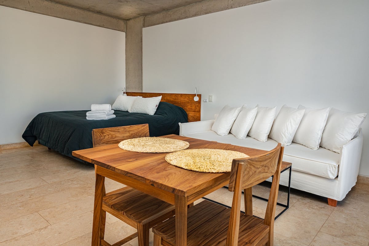 Una habitación con una mesa de madera, un sofá blanco y una cama con colcha verde oscuro.