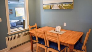 Un área de comedor con una mesa y sillas de madera, una ventana con vista a una pared azul y un sofá, junto con un cuadro de una escena de playa.