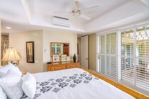 A hotel room with a bed, a ceiling fan, an air conditioning unit, a window with blinds, and a bedside table with a lamp and framed pictures.