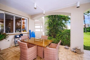 An outdoor patio with a glass-top table surrounded by wicker chairs, a window with a view, and a small garden area with a turtle.