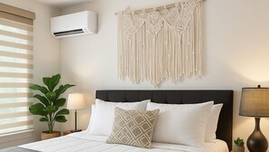 Un lit bien fait, avec une tête de lit sombre, une décoration murale en macramé et une plante en pot placée près d’une fenêtre munie de stores.