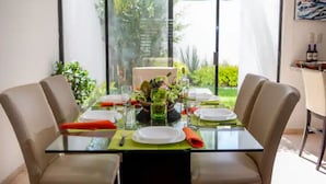 Une table à manger pour deux, avec un plateau en verre, entourée de chaises de couleur beige, et une vue sur la verdure grâce à de grandes fenêtres.