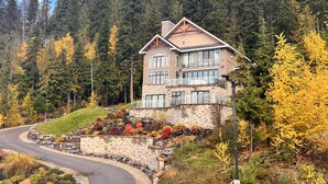 A two-story house with a stone facade, large windows, and a balcony overlooking a forested area. - RMR: Luxury Ski-in/out Mountain-side 4 Bedroom Home (Sleeps 10).  (Revelstoke)