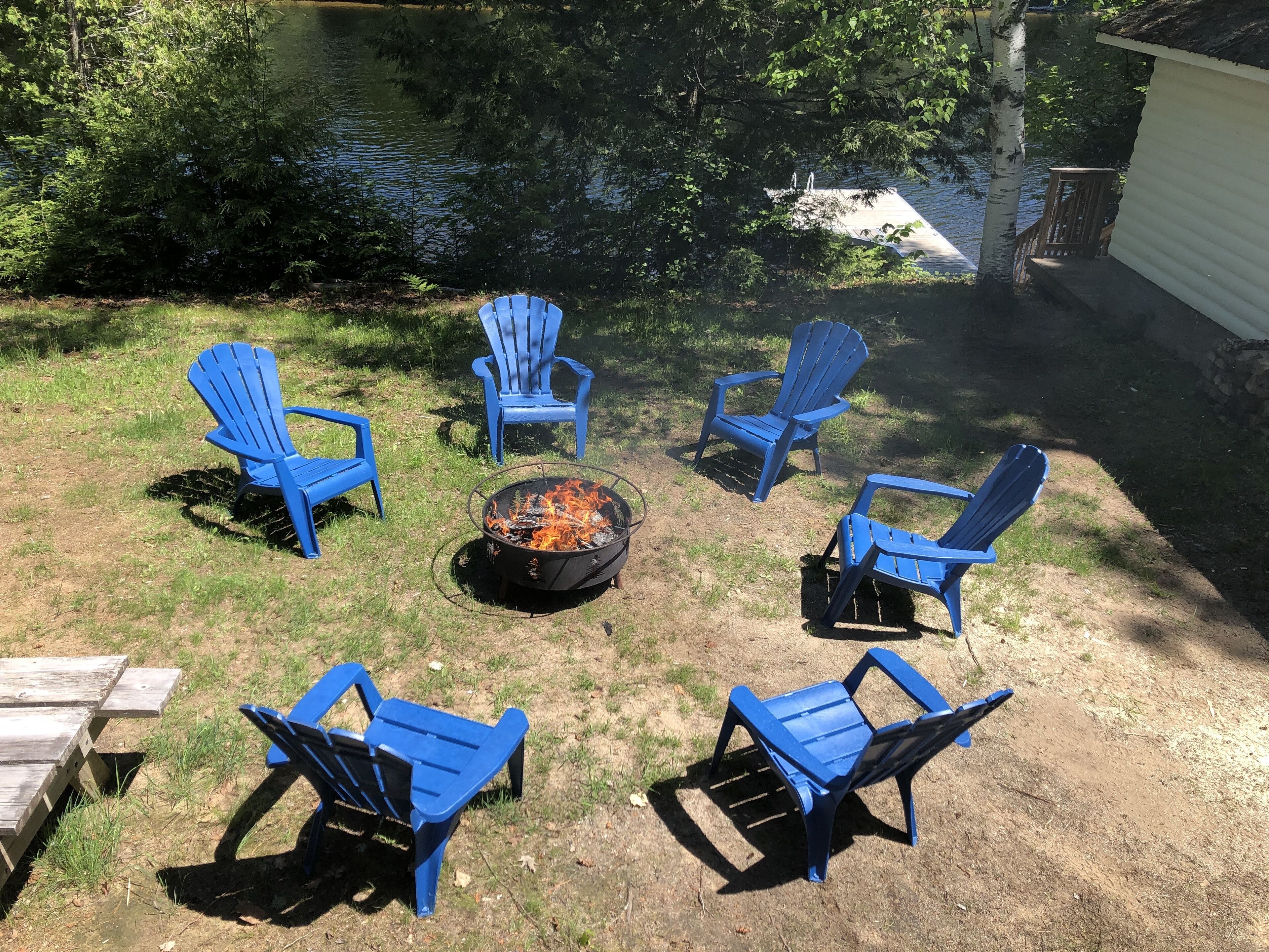 A backyard with a fire pit surrounded by six blue Adirondack chairs and a wooden deck.