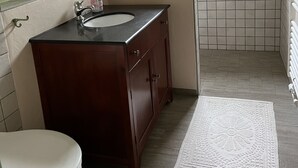 Un baño con tocador de madera, un lavamanos blanco, un inodoro, una ducha con puerta de vidrio y una ventana con diseños rojo y blanco.
