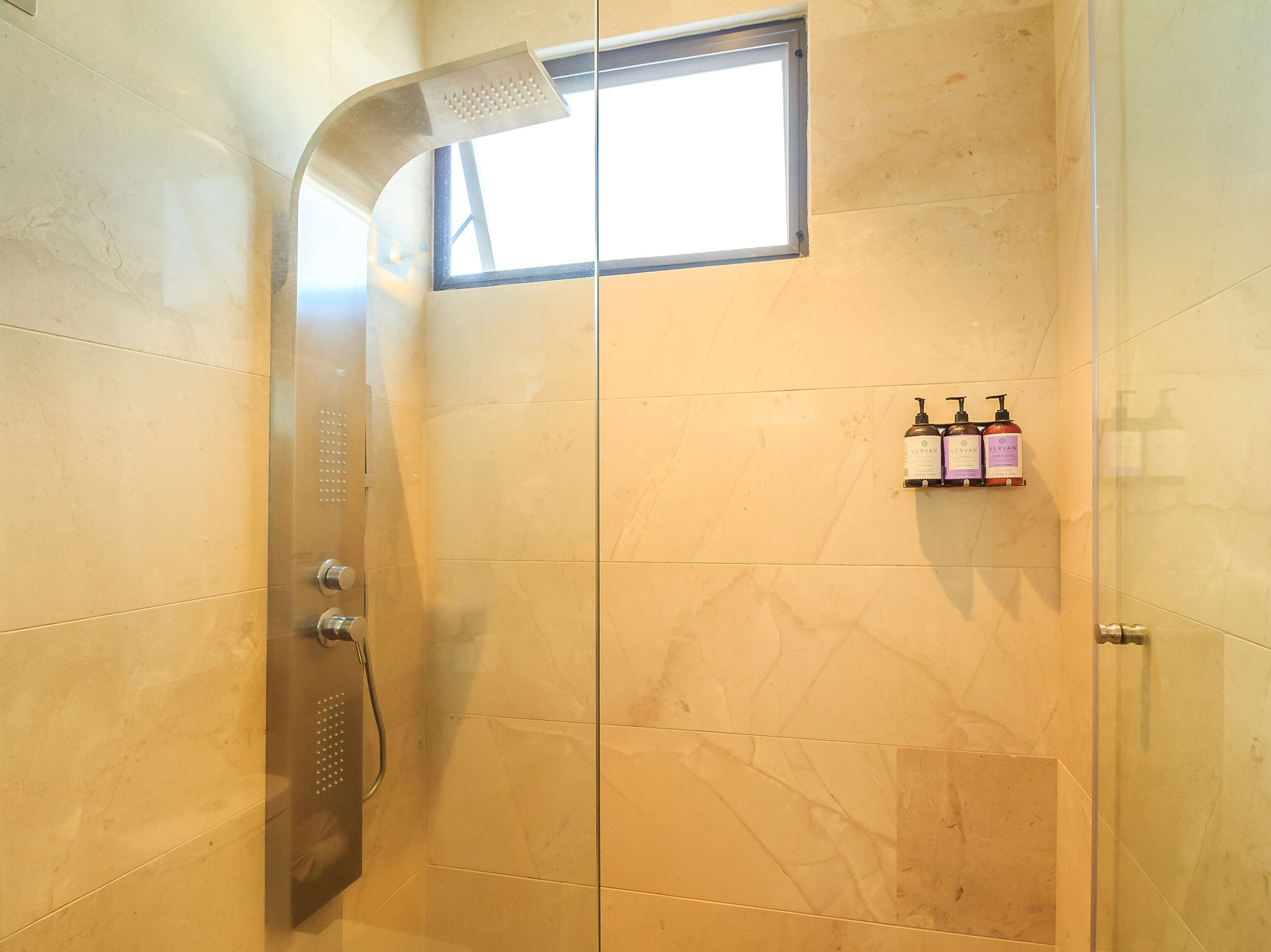 Ducha con cabezal de ducha curvo, una ventana pequeña y un estante con dos botellas de champú Vervan.