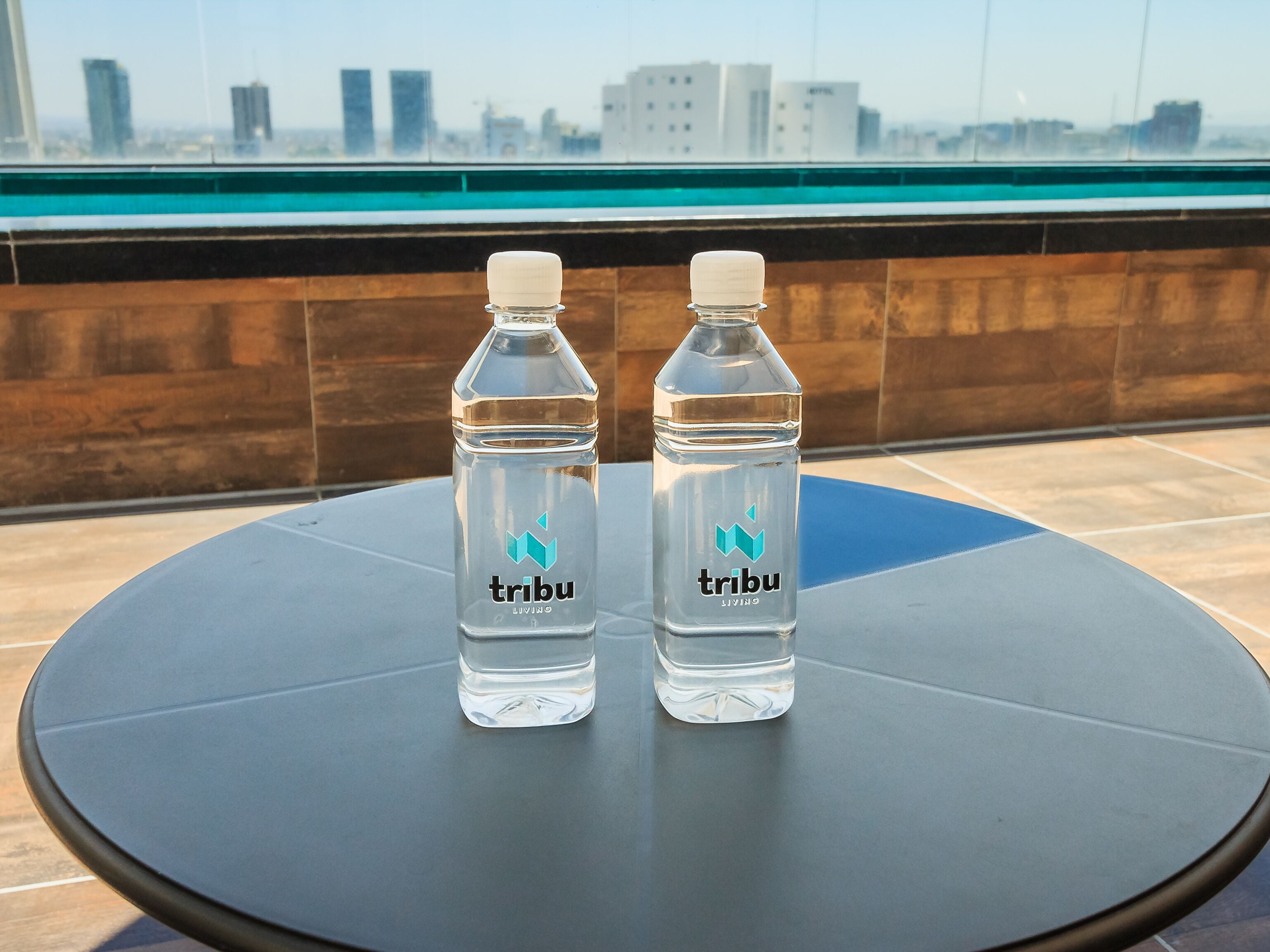 Deux bouteilles d’eau transparente, portant l’étiquette « tribu living », sont posées sur une table ronde devant laquelle on aperçoit un paysage urbain.