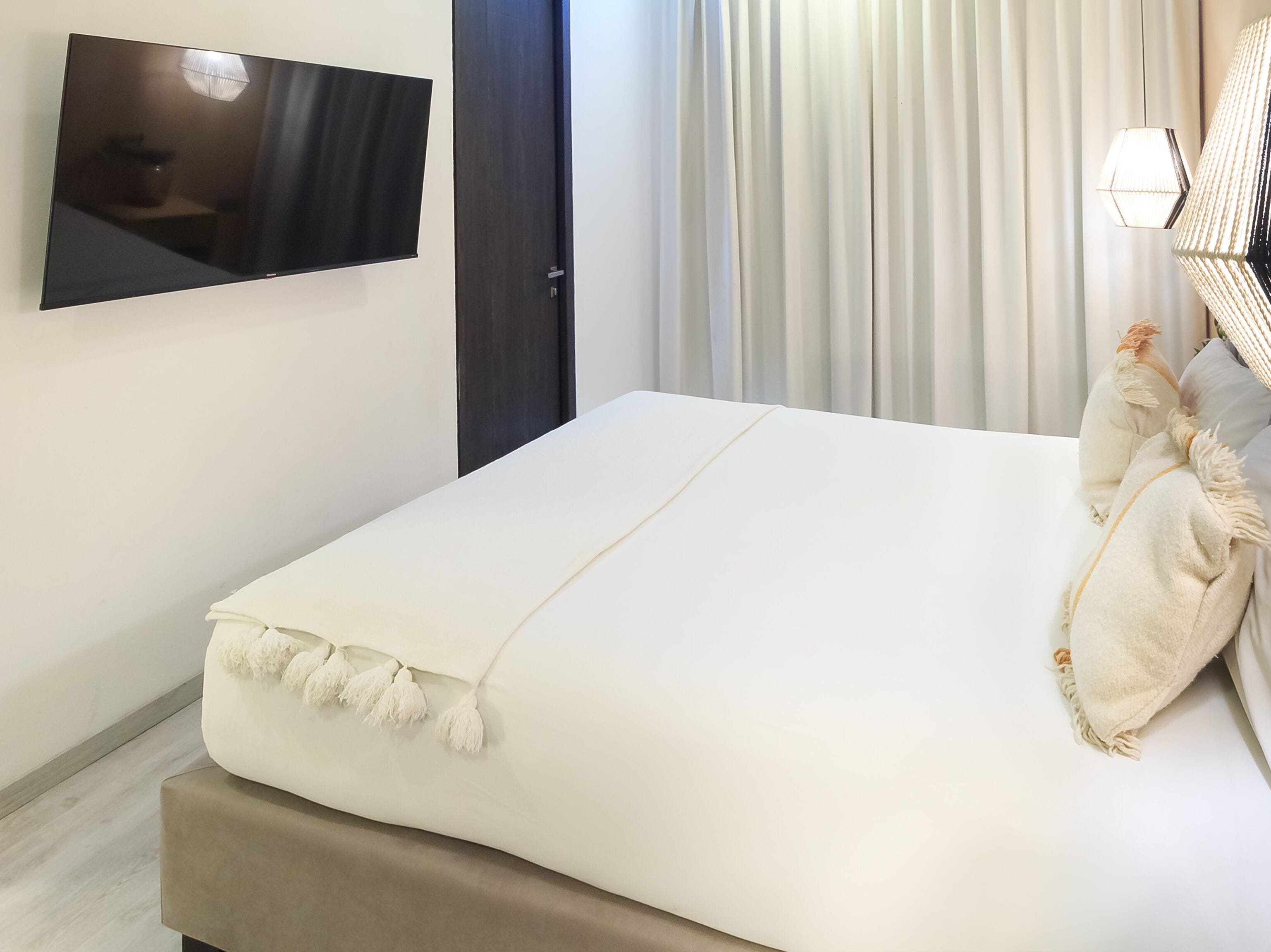 Una cama bien hecha con edredón blanco, un televisor de pantalla plana montado en la pared y una puerta con marco oscuro.