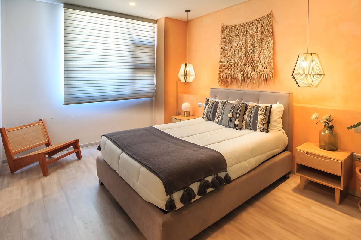 Une chambre moderne avec un grand lit, une table de chevet avec une lampe, un petit banc en bois et une fenêtre avec des stores.
