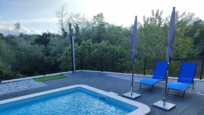 Zwembad met blauw water, omgeven door een betegeld terras. Twee blauwe ligbedden en twee parasols zijn aanwezig. Het terras wordt begrensd door een lage muur en een hek, met bomen en groen in de achtergrond.