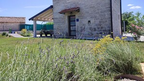 Een stenen gebouw met een pannendak, een overdekte patio en een tuin met lavendel en gele bloemen.