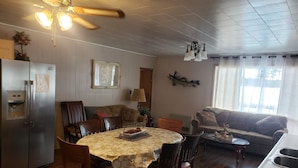 A dining area with a table set for four, surrounded by wooden chairs. A refrigerator is visible to the left, and a sofa is in the background.