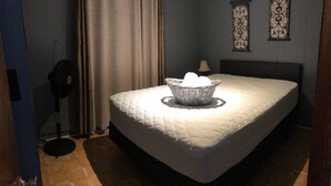 A bedroom with a bed, a standing fan, a wall-mounted lamp, and a decorative wall piece.