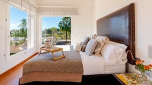 A bedroom with a large bed, a wooden bedside table, and a view of palm trees and buildings through the window.