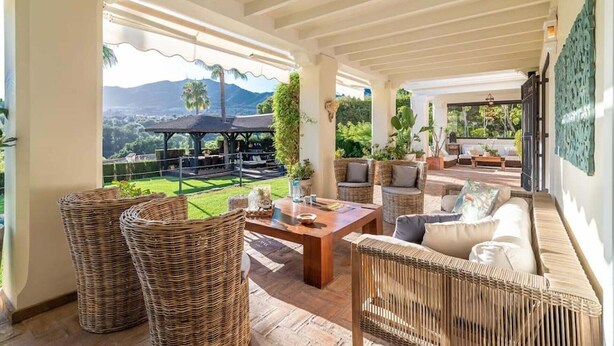 A covered outdoor seating area with wicker furniture, a wooden table, and a view of a mountainous landscape.