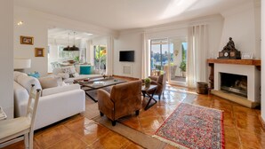 A spacious living room with a white sofa, a brown armchair, a coffee table, and a fireplace. There is a television mounted on the wall and a large window providing natural light.