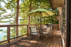 A wooden deck with outdoor furniture, a table, and an umbrella, overlooking a body of water and trees. - The Overlook | Luxury Lakeside Home with Dock (Harrison Hot Springs)