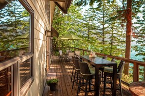 A wooden deck with outdoor seating, a table, and chairs, overlooking a forested area and a body of water. - The Overlook | Luxury Lakeside Home with Dock (Harrison Hot Springs)