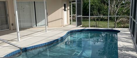 A covered outdoor swimming pool with a curved design, surrounded by a white fence and a house with a beige exterior.