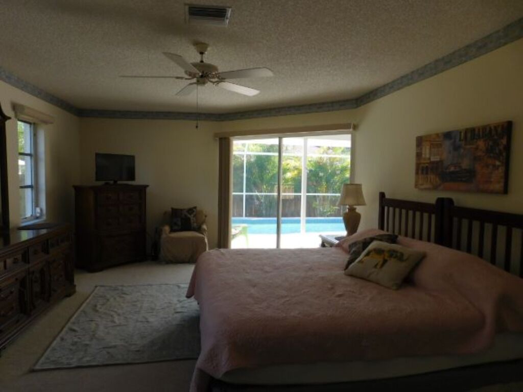 A bedroom with a bed, a ceiling fan, a dresser, a TV, a window with a view of greenery, and a painting on the wall.
