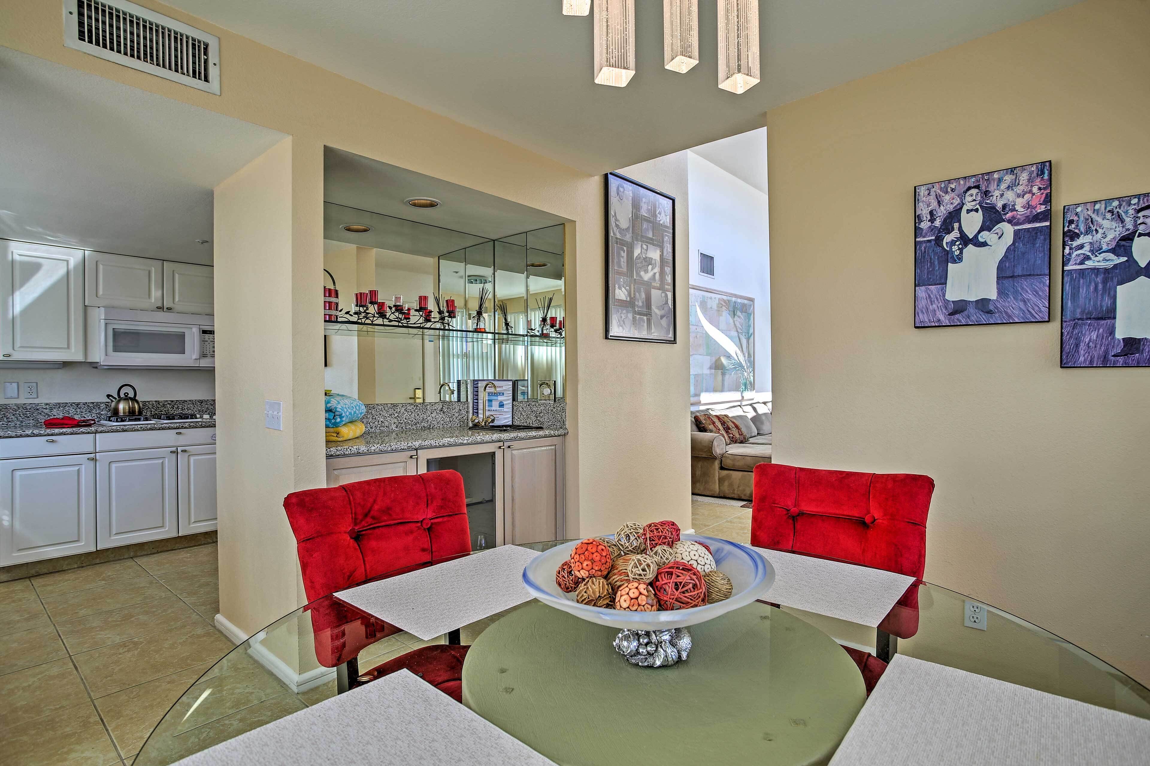 A dining area with a glass table, red chairs, and a bowl of decorative balls. A kitchen with white cabinets and a microwave is visible in the background.