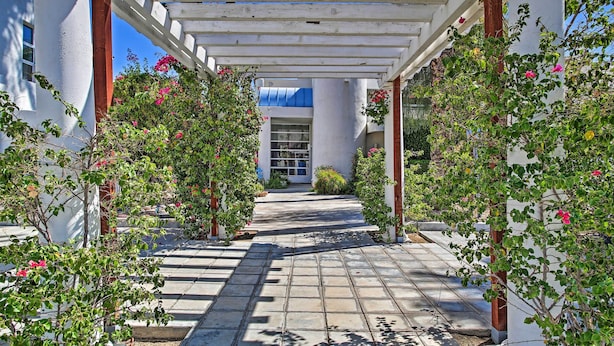A covered walkway with white columns and a tiled floor, surrounded by flowering plants.