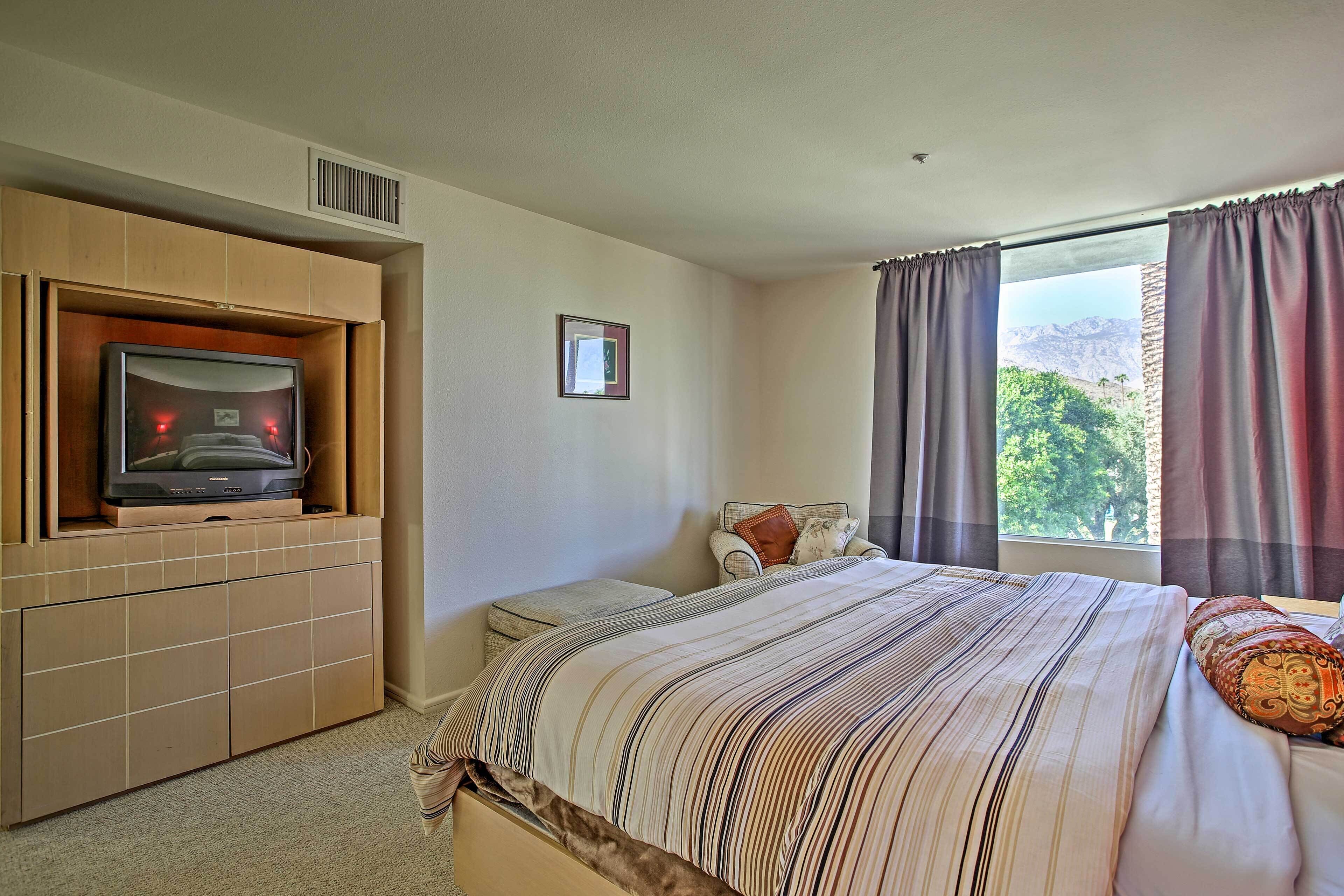 A bedroom with a bed, a television on a built-in shelf, a window with curtains, and a small bedside table.
