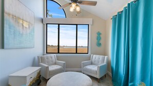 Une pièce avec deux fauteuils blancs, un pouf rond, une commode blanche dont la peinture s’écaillait, une grande fenêtre donnant sur une plage et un ventilateur de plafond.